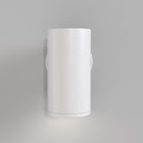 The FOCUS S Down Wall Light - White is a sleek, cylindrical fixture casting light both upward and downward. Mounted on a neutral gray wall, its white finish offers a minimalist aesthetic that enhances any modern industrial decor.