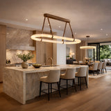 A modern kitchen with a large stone island, beige upholstered bar stools, wood cabinetry, and the Cintura LED Small Alabaster Floating Suspension Light – Soft White adding elegance above. The dining area features large windows with views of greenery.