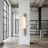 A modern minimal loft features large windows, concrete floors, exposed beams, and metal shelving. At the center, a Zarune LED Sculpted Curve Alabaster Wall Light in Matte Black & Soft White adds a striking sculptural touch on a white pillar.