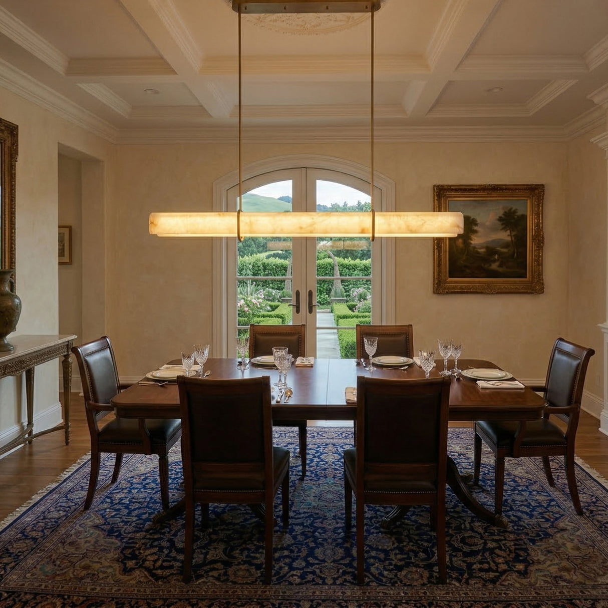 Niori Zarune LED linear alabaster chandelier in brushed brass, suspended above a mahogany dining table in a coffered-ceiling room with garden view.