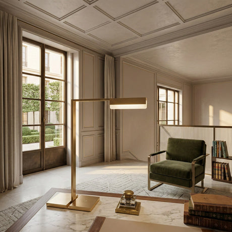 A bright, elegant living room with tall windows, beige walls, a green armchair, and bookshelves. The Solin LED Alabaster Table Lamp in brushed brass sits on a marble table, its warm light highlighting the cozy, sophisticated decor.