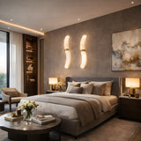 Modern bedroom with a large bed, neutral bedding, two Aureli LED Curved Alabaster Wall Lights in soft white and brushed brass, abstract art, bedside tables with lamps, a round coffee table with flowers, and cozy seating by floor-to-ceiling windows.