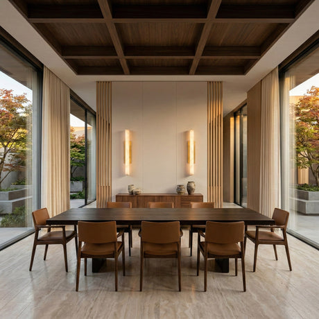 Modern dining room with a dark wood table, eight brown chairs, large windows, marble floor, minimalist decor, light wood ceiling accents, and ambient accent lighting featuring the Selvara LED Medium Alabaster Wall Light in brushed brass. Trees outside.