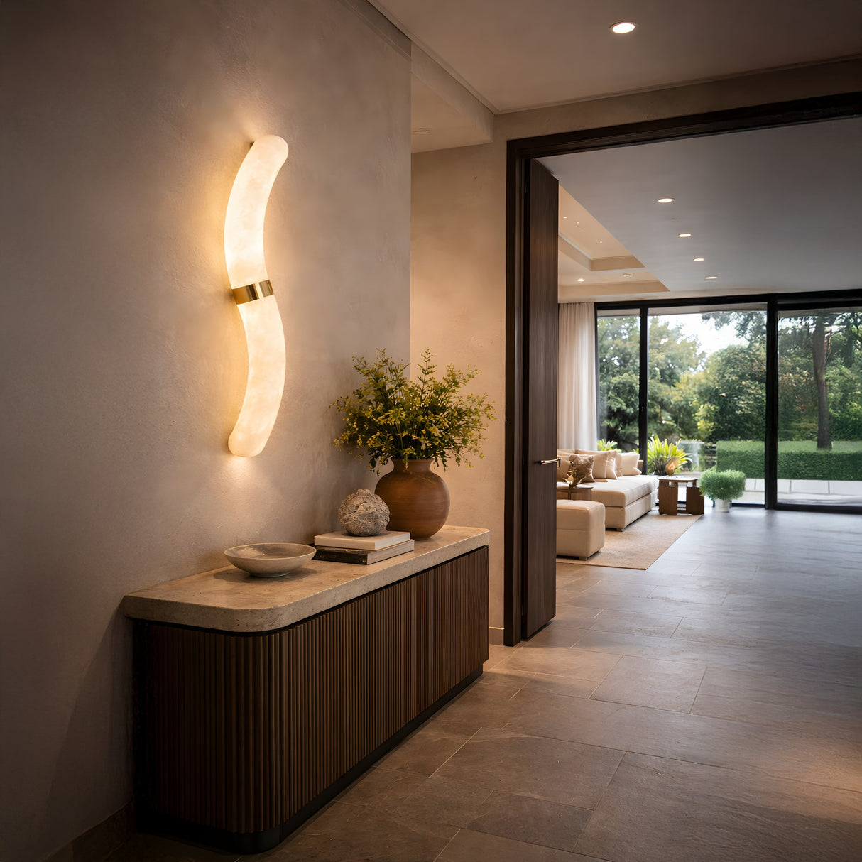 A modern entryway features the Aureli LED Curved Alabaster Wall Light in Soft White & Brushed Brass above a console table with vases and a bowl. The living room in the background has a sofa and large windows overlooking a green yard.
