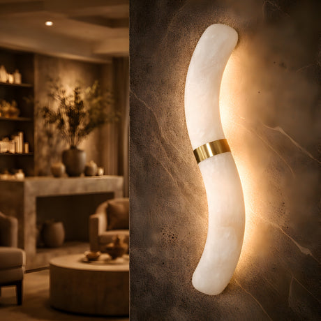 The Aureli LED Curved Alabaster Wall Light in soft white with a brushed brass accent emits warm light and is ideal for mounting on textured brown walls in an elegant, softly lit living room.