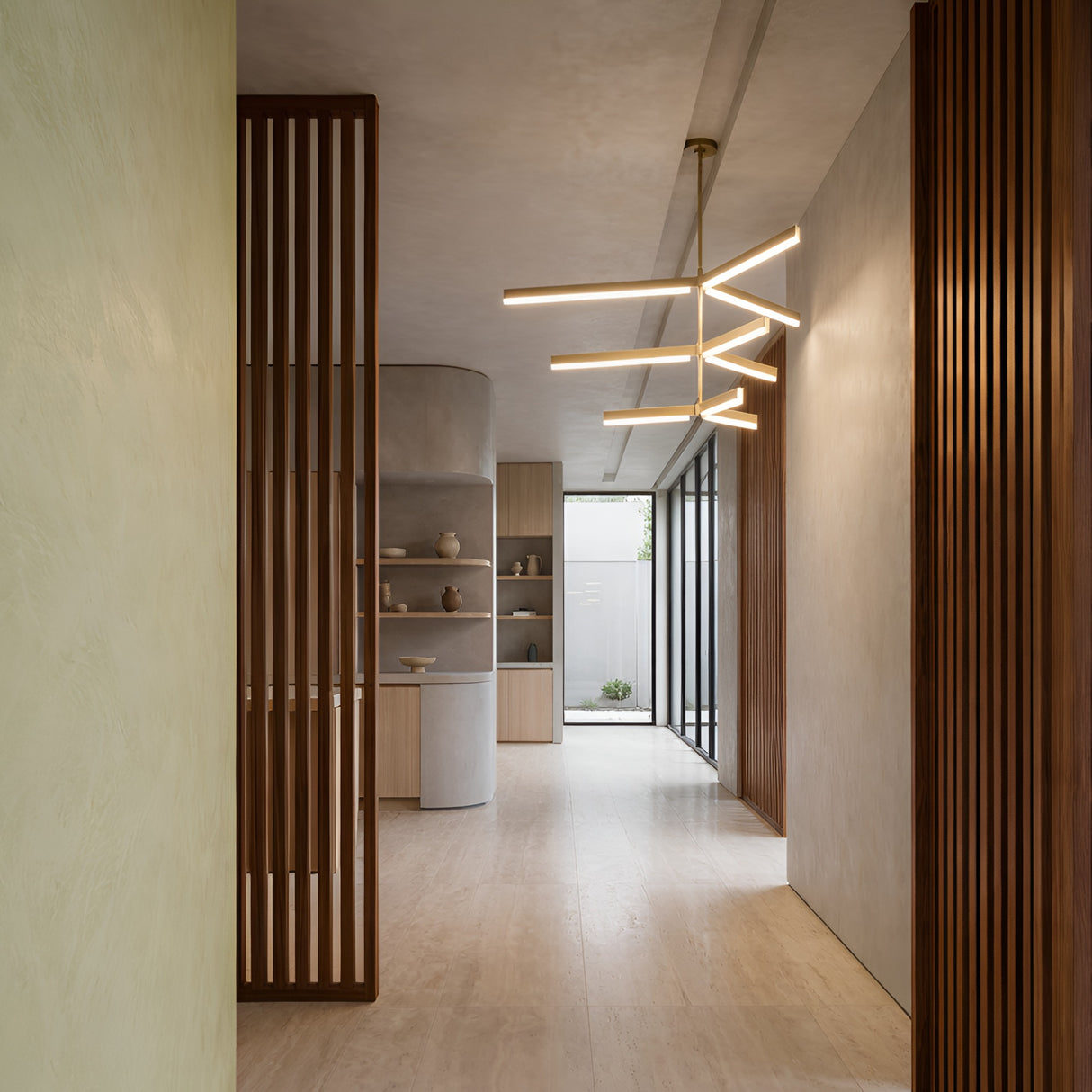 A minimalist hallway with light wood floors, vertical wooden slats, neutral walls, and pottery on shelves is elegantly lit by the Elvaris LED 3 Tier Radial Alabaster Chandelier in brushed brass, while large windows at the end bring in natural light.