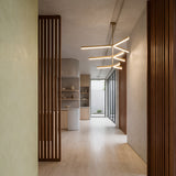 A minimalist hallway with light wood floors, vertical wooden slats, neutral walls, and pottery on shelves is elegantly lit by the Elvaris LED 3 Tier Radial Alabaster Chandelier in brushed brass, while large windows at the end bring in natural light.