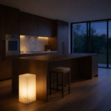 A modern kitchen at dusk with dark wooden cabinets, marble backsplash, a built-in oven, and a large island. In the foreground sits the Monvara 1 Light Small Monolith Alabaster Floor Lamp in soft white beside a stool; trees show through large windows.