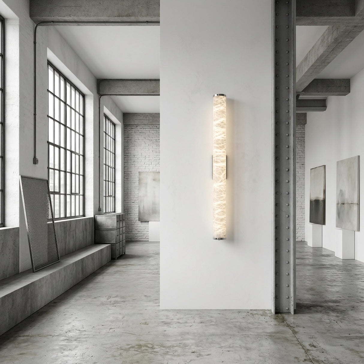 A modern, industrial-style art gallery with large windows, concrete floors, and abstract paintings on white walls features the Corvane LED Medium Slim Cylindrical Alabaster Wall Light - Soft White illuminated on a central wall.
