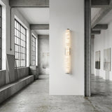 A modern, industrial-style art gallery with large windows, concrete floors, and abstract paintings on white walls features the Corvane LED Medium Slim Cylindrical Alabaster Wall Light - Soft White illuminated on a central wall.