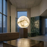 A modern interior features the Orlena 3 Light Globe Alabaster Floor Lamp - Soft White on a wooden pedestal, surrounded by tall windows, green marble walls, and wooden furniture, creating a luxurious and serene atmosphere.