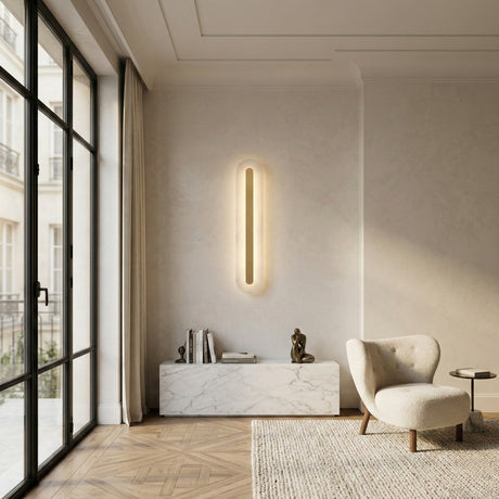 A modern living room with large windows, a marble cabinet with books and sculpture, a cozy cream chair on herringbone wood floors, and an Averra LED Large Round Alabaster Wall Light (Soft White & Brass) adding minimalist elegance.