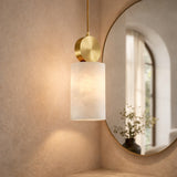 The Serano 1 Light Large Vertical Pillar Alabaster Single Pendant Light in soft white and brass hangs beside a round mirror on a beige wall, reflecting a window and a vase with greenery.