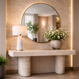 A modern entryway with a round mirror above a stone console, featuring the Virello 3 Light Medium Compact Alabaster Table Lamp in soft white and a vase of white flowers. Neutral walls and a potted plant finish the elegant, minimalist look.