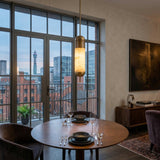 A round dining table for two with black plates and wine glasses sits by large windows overlooking the city, while the Zerith LED Fluted Tube Alabaster Single Pendant Light in soft white & brushed brass adds modern elegance overhead.