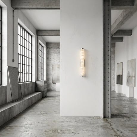 A spacious, minimalist art gallery with large windows and white walls displays abstract paintings illuminated by the Corvane LED Small Slim Cylindrical Alabaster Wall Light - Soft White, emitting a soft, elegant glow.