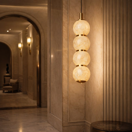 A modern hallway with marble walls and floors features the Lumina 4 Light Orb Alabaster Single Pendant Light in soft white and brass—four glowing spheres stacked vertically. Wall sconces and an archway are visible in the background.