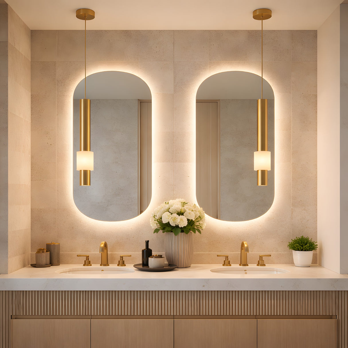 Modern bathroom with double sinks, gold fixtures, two oval backlit mirrors, and the Solenne LED Alabaster Single Pendant Light in Soft White & Brushed Brass. White flowers, soap dispensers, and a green potted plant sit on the light stone countertop.