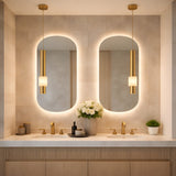 Modern bathroom with double sinks, gold fixtures, two oval backlit mirrors, and the Solenne LED Alabaster Single Pendant Light in Soft White & Brushed Brass. White flowers, soap dispensers, and a green potted plant sit on the light stone countertop.