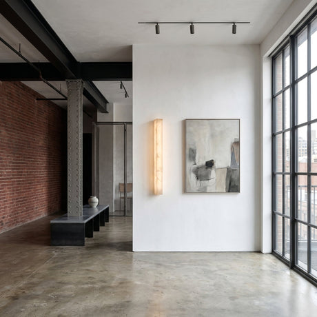 A modern industrial loft with exposed brick, metal beams, polished concrete floor, large grid windows, a minimalist bench, ceramic vase, abstract wall art, and the Valmera LED Small Slim Vertical Alabaster Wall Light in soft white illuminating a white wall.