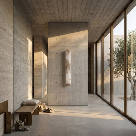 A minimalist, sunlit room with concrete walls and floor features a floating stone sculpture, wooden benches, scattered rocks, and the Arvella LED Small Column Alabaster Wall Light in soft white, with large windows revealing an olive tree outside.