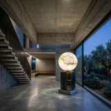 A modern concrete interior features a floating staircase, floor-to-ceiling windows with garden views, and the Orlena 3 Light Globe Alabaster Floor Lamp - Soft White beside a glowing spherical sculpture, creating a serene minimalist vibe.