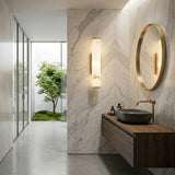 Modern bathroom featuring marble walls, a round gold-framed mirror, stone vessel sink on wooden vanity, gold faucet, Montavo LED Large Tube Alabaster Wall Light in soft white & brushed brass, and a window view of mossy greenery.