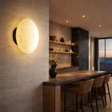 A modern kitchen with a stone accent wall features the Solenne LED Small Halo Alabaster Wall Light in soft white and matte black, wooden shelves with pottery, a long wooden island with woven barstools, and large windows revealing a sunset view.