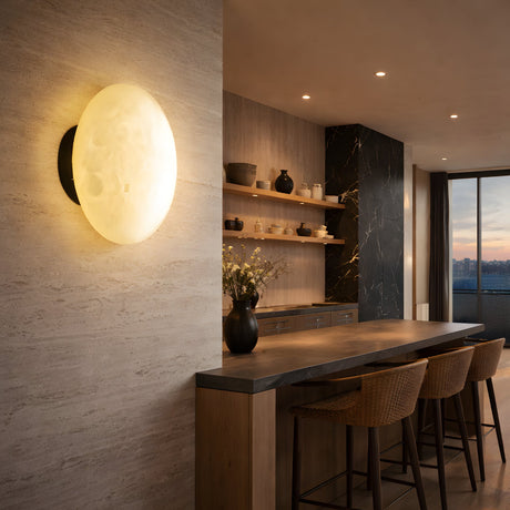 A modern kitchen with a stone accent wall features the Solenne LED Small Halo Alabaster Wall Light in soft white and matte black, wooden shelves with pottery, a long wooden island with woven barstools, and large windows revealing a sunset view.