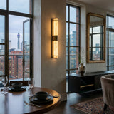 A modern dining area with a round table for two, city-view windows, illuminated by the Averna LED Large Framed Alabaster Wall Light in Soft White & Brushed Brass, plus a sideboard with a vase and a large mirror reflecting the room.