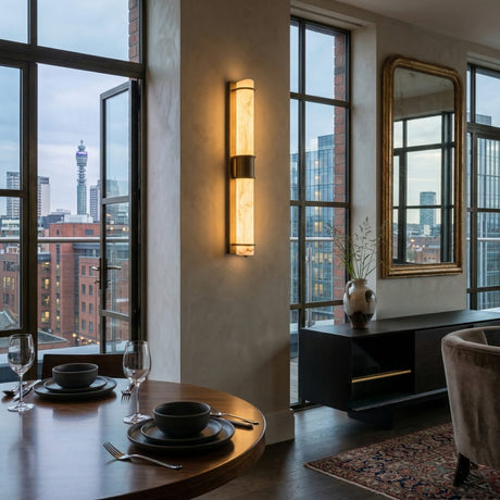 A modern dining area with a round table for two, city-view windows, illuminated by the Averna LED Large Framed Alabaster Wall Light in Soft White & Brushed Brass, plus a sideboard with a vase and a large mirror reflecting the room.