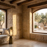 A cozy stone-walled room features large windows, a cushioned window seat, and a small round table topped with the Veloris LED Carved Alabaster Table Lamp in Brushed Brass & Soft White. Sunlight streams in, illuminating the patio with chairs and trees outside.