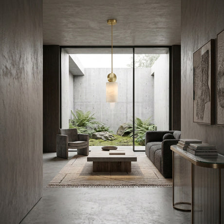 A minimalist living room with gray tones, modern furniture, and abstract art is softly illuminated by the Serano 1 Light Large Vertical Pillar Alabaster Pendant in soft white & brass. A large window reveals a tranquil rock and fern garden, filling the space with natural light.
