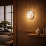 A modern Japanese-style room features the Orvani LED Large Oval Alabaster Wall Light - Soft White on wood paneling, a bonsai by shoji windows, and a wooden shelf with a ceramic vase and books. Warm lighting creates a cozy atmosphere.