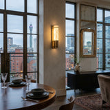 A modern dining area with a set table overlooks tall city buildings through large windows. The interior features the Averna LED Small Framed Alabaster Wall Light in Soft White & Brushed Brass, a sideboard with a vase, and a mirror.