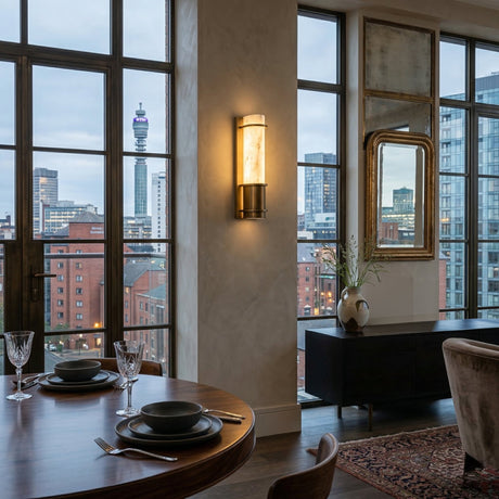 A modern dining area with a set table overlooks tall city buildings through large windows. The interior features the Averna LED Small Framed Alabaster Wall Light in Soft White & Brushed Brass, a sideboard with a vase, and a mirror.