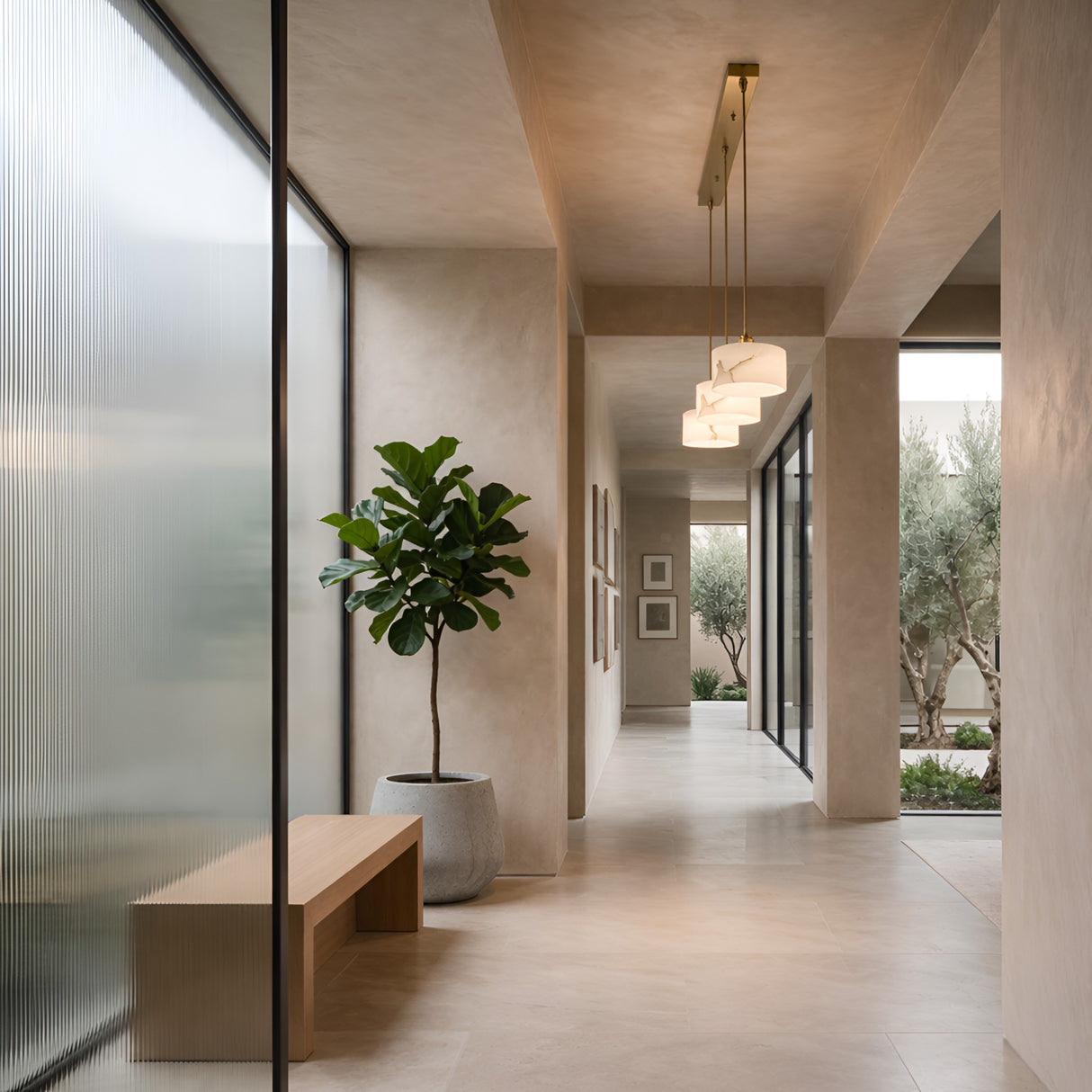 A modern hallway with neutral tones features a large potted plant, a wooden bench, and the Brinex 3 Light Linear Alabaster Bar Pendant Light in soft white & brushed brass. Floor-to-ceiling windows add brightness to this minimalist space.