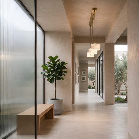 A modern hallway with neutral tones features a large potted plant, a wooden bench, and the Brinex 3 Light Linear Alabaster Bar Pendant Light in soft white & brushed brass. Floor-to-ceiling windows add brightness to this minimalist space.