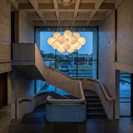 A modern concrete staircase with curved railings sits beneath the Auriel 19 Light Alabaster Chandelier in Soft White & Brushed Brass, its alabaster spheres glowing above a window overlooking landscaped gardens and a reflecting pool at dusk.