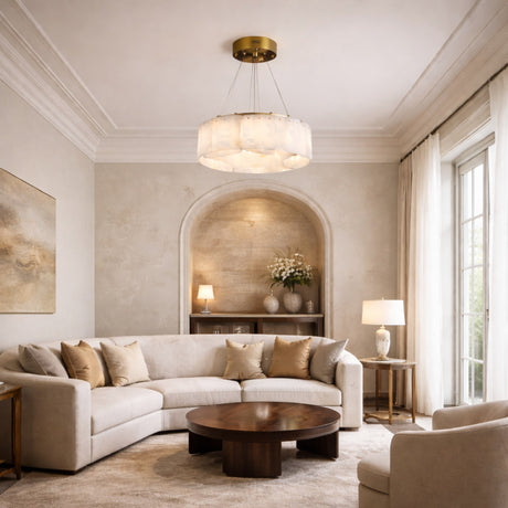 Elegant living room with a curved beige sofa, wooden coffee table, neutral-toned decor, large window with sheer curtains, and the Caldrisa 10 Light Layer Panel Alabaster Chandelier in Soft White & Brass. Niche shelves display vases and a lamp for cozy sophistication.