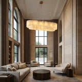 A spacious modern living room with tall windows, beige walls, and neutral furniture centers on the Arvello 12 Light Layered Panel Alabaster Linear Chandelier in Soft White & Brass. Two sofas and armchairs encircle a round coffee table on a gray rug.