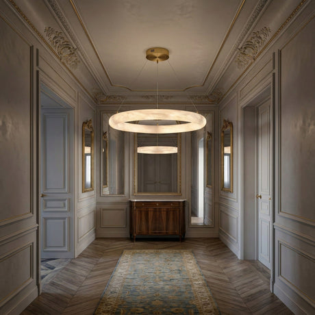 A narrow, elegant hallway features ornate molding, gold accents, tall mirrors, and two Seraphis LED Small Alabaster Chandeliers in Soft White illuminating a wooden console and patterned rug on herringbone wood floors.
