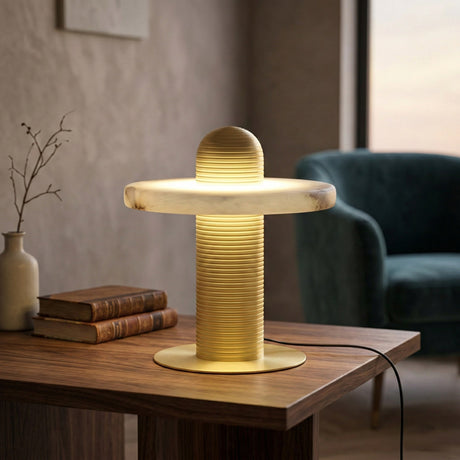 The Nyxara LED Single Tiered Alabaster Table Lamp in soft white and brass sits on a wooden side table beside stacked books and a ceramic vase, with a green armchair and window in the background.