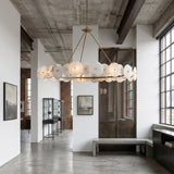 A modern industrial-style room with large windows, white walls, and an exposed concrete ceiling features the Solaryn LED Linear Disc Alabaster Chandelier in soft white & brass above a minimalist bench. Abstract art decorates the walls.