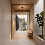 A modern hallway with light wood and stone accents features a built-in bench, a potted plant, and the Arvion 1 Light Slim Capsule Alabaster Pendant in brushed brass, overlooking a minimalist garden through a large glass window.