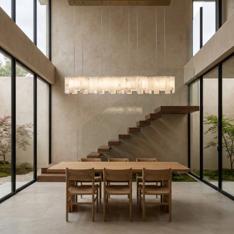 A modern dining room with tall ceilings, large windows showing greenery, a wooden table for six, floating staircase, stone geometry accents, and the Caldrisa 8 Light Layer Panel Alabaster Bar Pendant Light in soft white & brass.