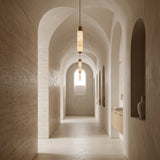 A minimalist hallway with arched ceilings, beige walls, and vases in built-in alcoves features the Elviro LED Large Slim Tube Alabaster Single Pendant Light in soft white and brushed brass. Natural light from a small end window adds to the serene, modern vibe.