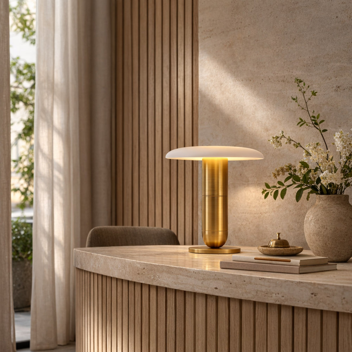 A modern interior showcases a beige stone desk with the Luxelle LED Single Tiered Alabaster Disc Table Lamp in soft white and brass, a flower vase, and books. Vertical wooden panels and sheer curtains enhance the softly lit, minimalist space.