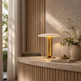 A modern interior showcases a beige stone desk with the Luxelle LED Single Tiered Alabaster Disc Table Lamp in soft white and brass, a flower vase, and books. Vertical wooden panels and sheer curtains enhance the softly lit, minimalist space.