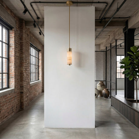 A modern industrial interior with exposed brick, ceiling pipes, large windows, concrete floors, and decorative pottery features a white partition adorned by the Elviro LED Small Slim Tube Alabaster Single Pendant Light in soft white and brushed brass.